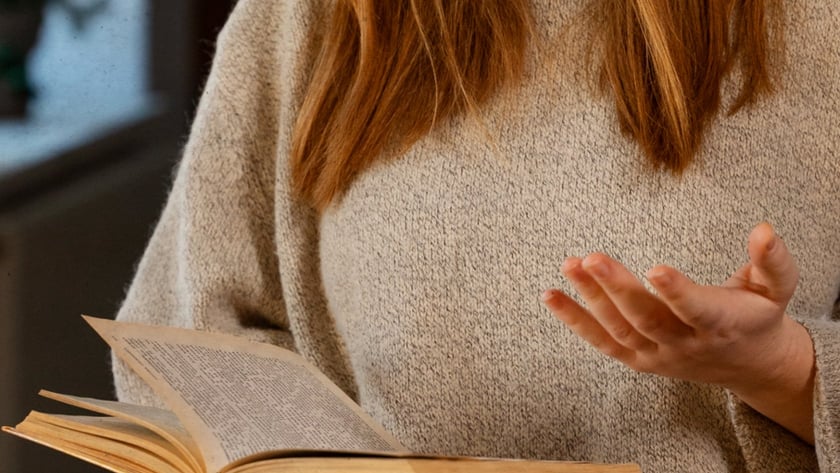 Mulher de cabelo ruivo e suéter bege segura um livro antigo aberto e gesticula.