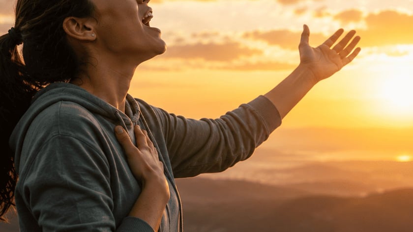 Mulher de capuz e rabo de cavalo, de olhos fechados, canta com a mão no peito ao pôr do sol.
