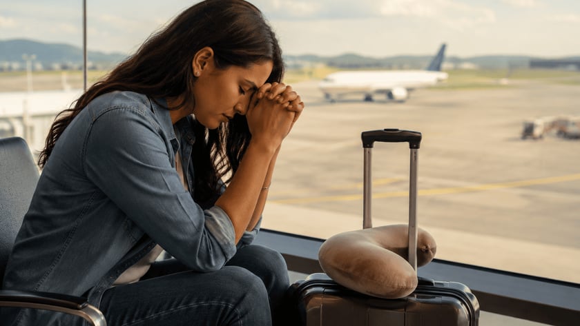 Mulher de cabelo escuro, com as mãos juntas na testa, sentada em aeroporto ao lado de mala.