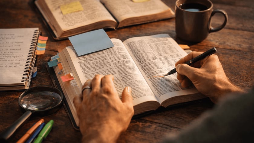 Mão de uma pessoa sobre livro aberto, com caneta, cadernos, lupa e post-its na mesa.