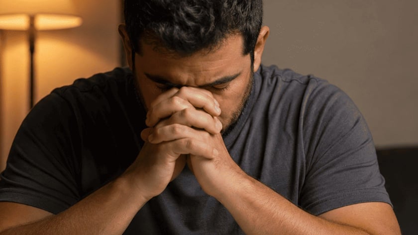 Homem de camiseta cinza, de cabeça baixa e mãos unidas, diante de uma Bíblia aberta.