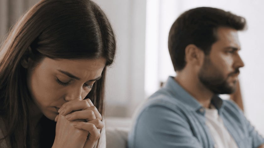 Mulher de cabeça baixa e mãos juntas, ao lado de homem de braços cruzados no sofá.