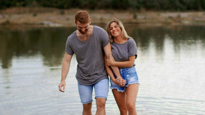 Casal de camisetas cinza e shorts jeans caminha de braços dados em um lago raso.