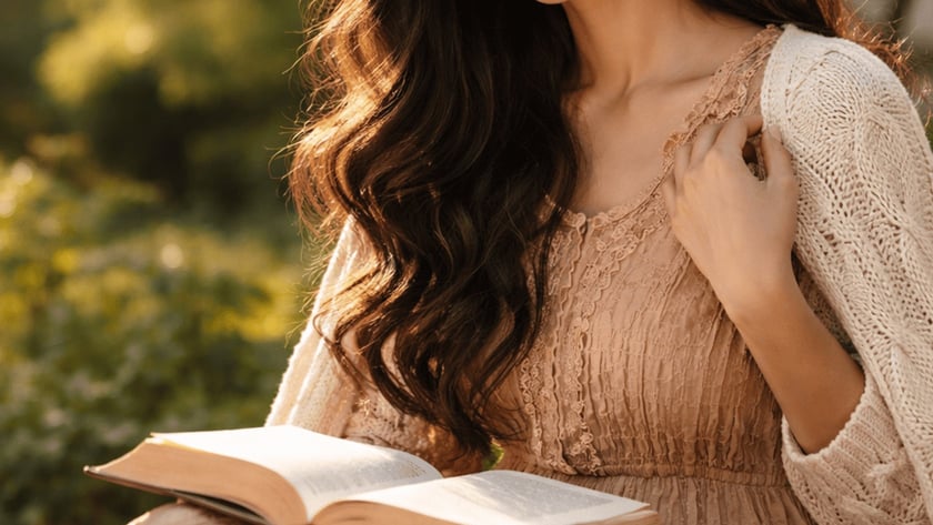 Mulher de cabelos longos e ondulados segura um livro aberto no colo, sentada ao ar livre.