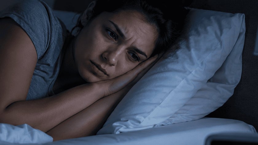 Mulher deitada na cama, de olhos abertos, olhando para o despertador na mesa de cabeceira.