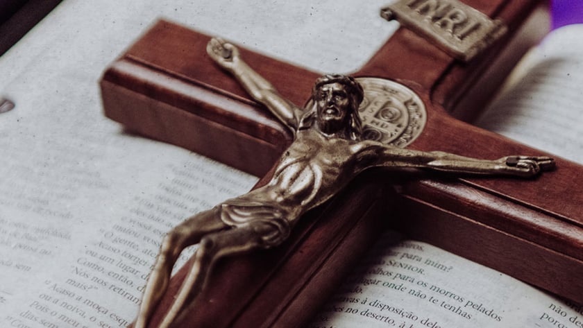 Crucifixo de madeira com Cristo metálico sobre Bíblia aberta, com tecido roxo ao fundo. "INRI"