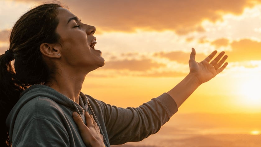 Mulher de capuz e rabo de cavalo, de olhos fechados, canta com a mão no peito ao pôr do sol.