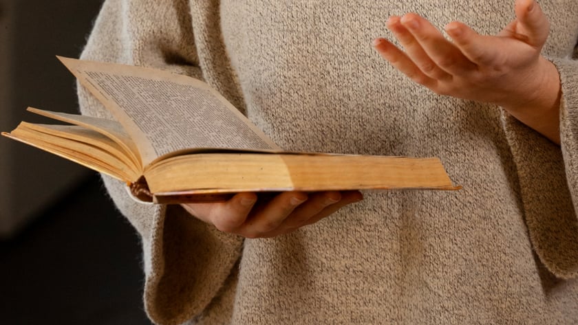 Mulher de cabelo ruivo e suéter bege segura um livro antigo aberto e gesticula.
