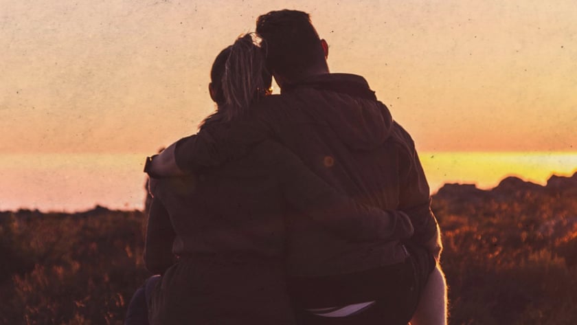 Casal abraçado, sentado de costas sobre uma pedra, olhando o horizonte ao pôr do sol.
