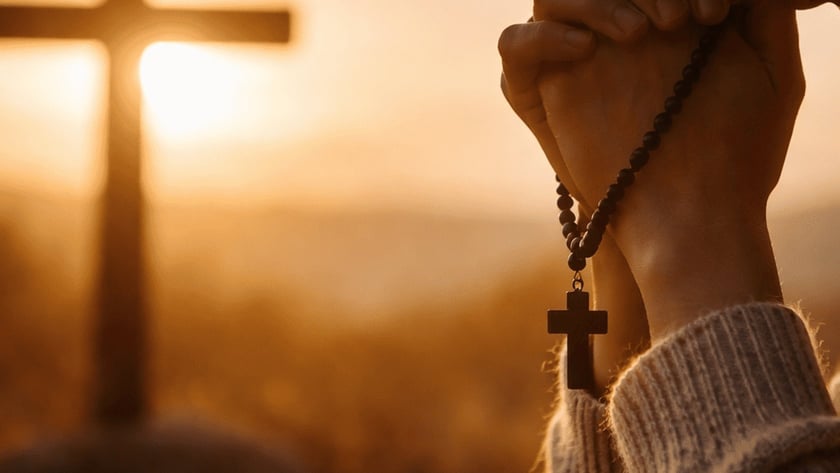 Mulher de suéter claro, de cabeça baixa, reza com um terço nas mãos diante de uma cruz.
