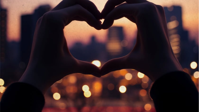Duas mãos formam um coração diante de uma cidade ao entardecer, com luzes desfocadas.
