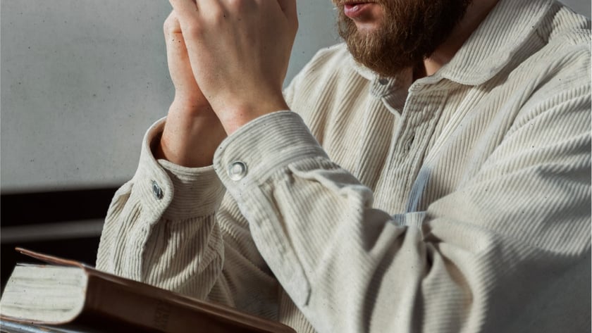 Homem barbudo de camisa bege, de olhos fechados e mãos unidas, ao lado de um livro.