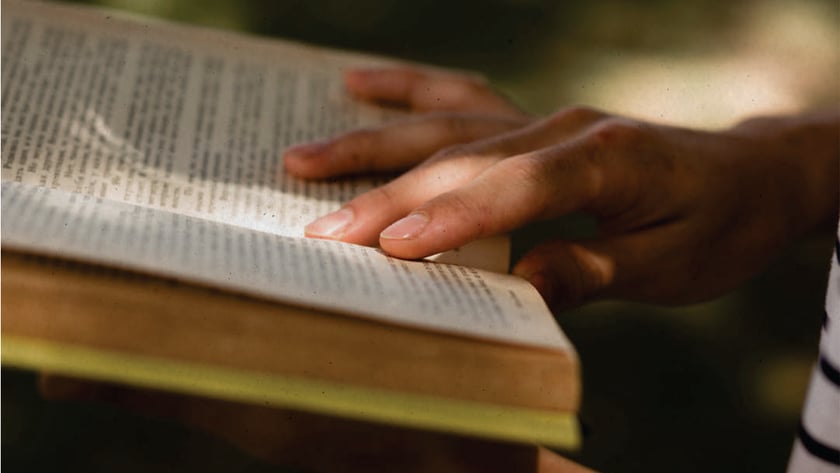 Mãos de uma pessoa segurando um livro aberto ao ar livre, com os dedos sobre a página.