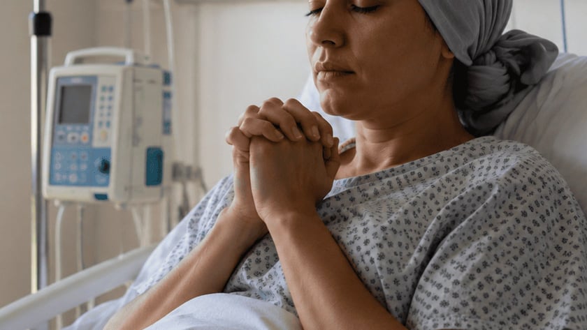 Mulher deitada em cama de hospital, de mãos juntas e olhos fechados, ao lado de soro.