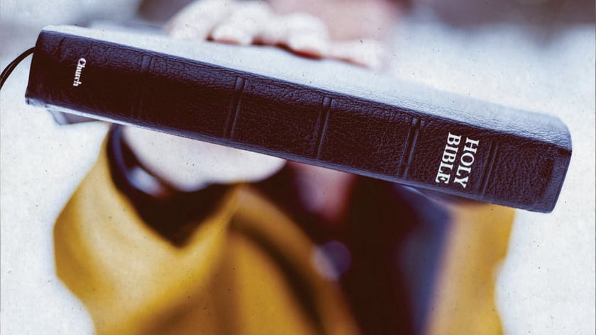 Pessoa de casaco amarelo estende uma Bíblia preta de capa de couro: "HOLY BIBLE".