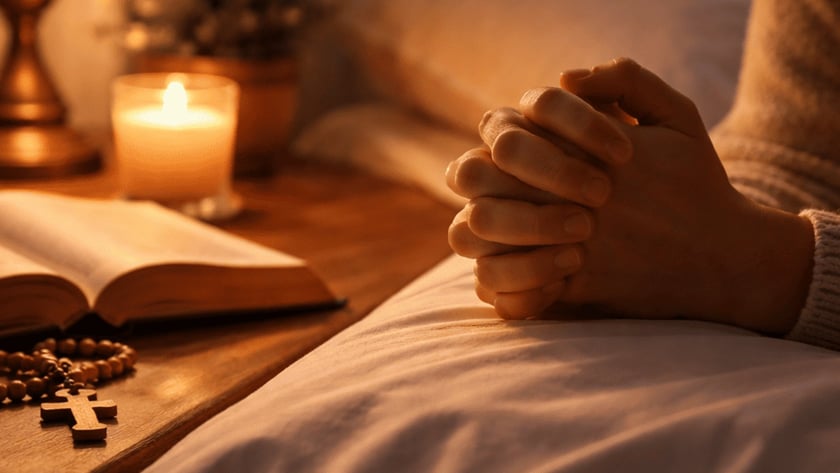 Mãos postas sobre a cama, ao lado de mesa com Bíblia aberta, terço e vela.