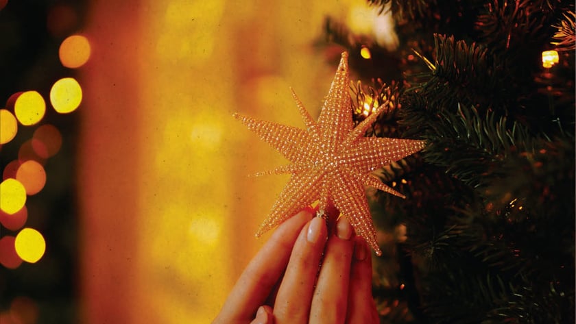 Duas mãos seguram uma estrela dourada de contas junto a uma árvore de Natal.