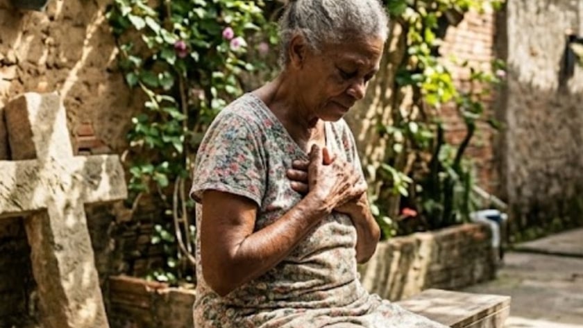 Idosa de vestido florido sentada num banco de madeira, com as mãos sobre o peito, em um quintal.