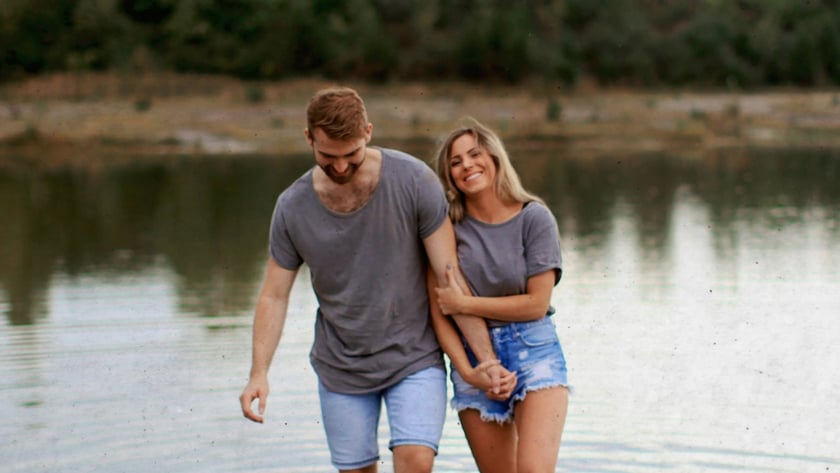 Casal de camisetas cinza e shorts jeans caminha de braços dados em um lago raso.