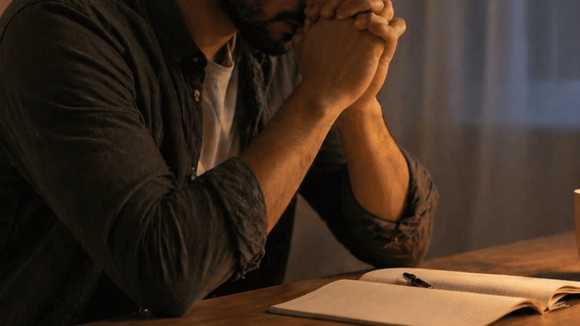Homem de barba com as mãos unidas junto ao rosto, sentado à mesa com caderno.