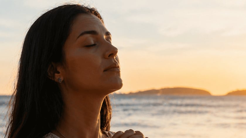 Mulher de cabelos escuros e olhos fechados, com as mãos unidas, na praia.