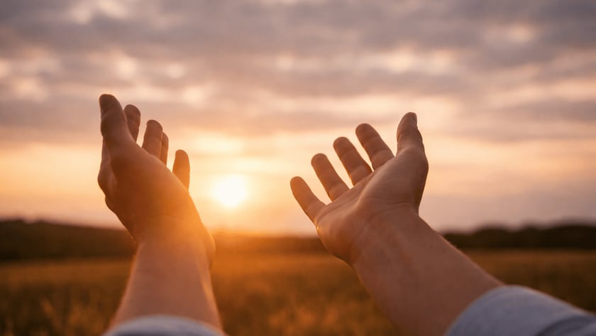 Duas mãos abertas erguidas em direção ao sol poente sobre um campo.