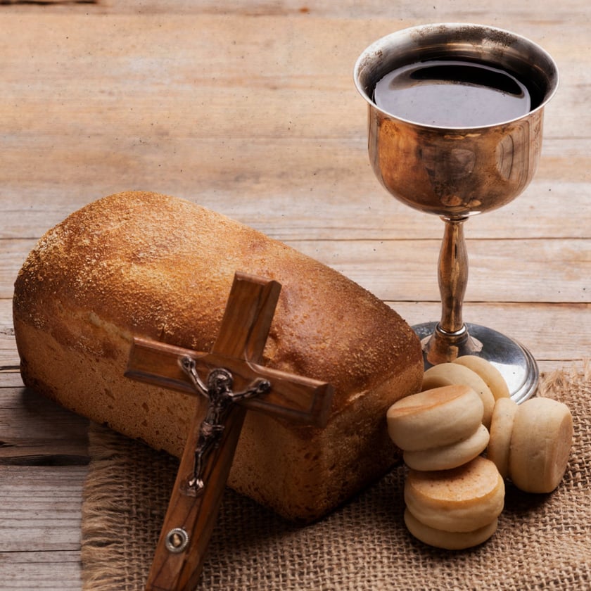 Crucifixo de madeira apoiado em pão de forma, ao lado de cálice com vinho e pãezinhos.