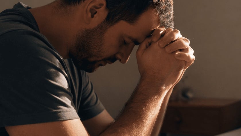 Homem de camiseta cinza, de olhos fechados e mãos postas na testa, ao lado de uma cama.