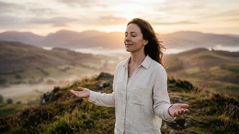 Mulher de olhos fechados e braços abertos em um morro com montanhas ao fundo.