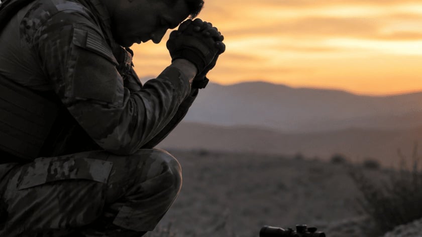 Soldado ajoelhado de cabeça baixa e mãos unidas, com capacete e fuzil no chão, ao pôr do sol.