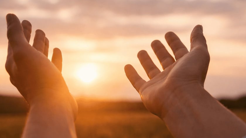 Duas mãos abertas erguidas em direção ao sol poente sobre um campo.