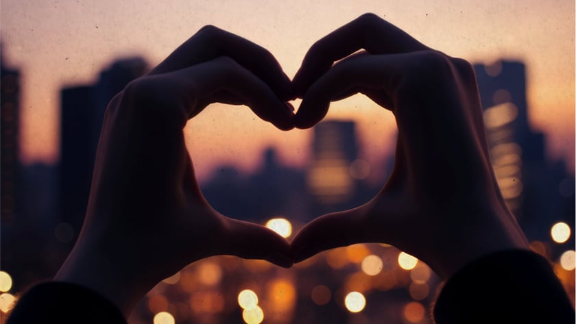 Duas mãos formam um coração diante de uma cidade ao entardecer, com luzes desfocadas.