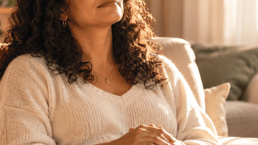 Mulher de cabelo cacheado, de olhos fechados, sentada no sofá com uma Bíblia aberta no colo.