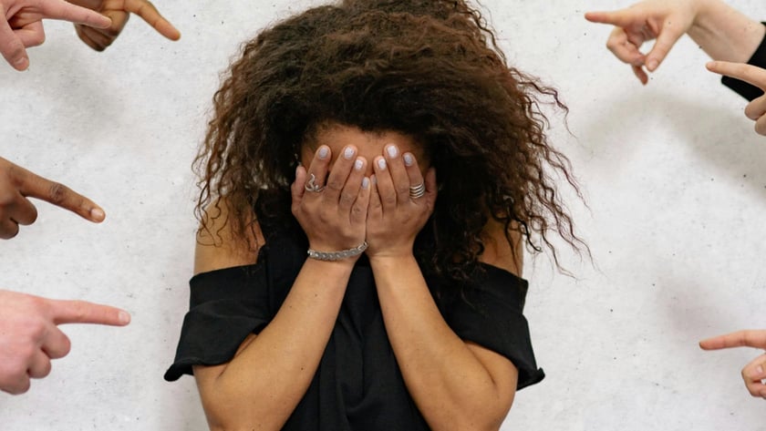 Mulher de cabelo cacheado cobre o rosto com as mãos, cercada por dedos apontados.