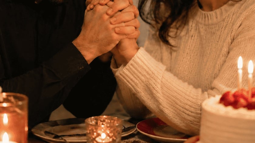 Casal de mãos dadas e cabeça baixa à mesa, com bolo de velas acesas.