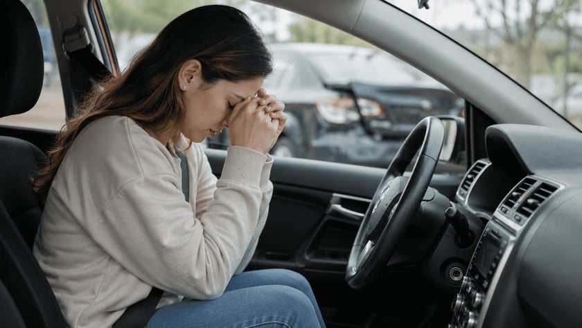 Mulher sentada ao volante de um carro, com o rosto apoiado nas mãos unidas.