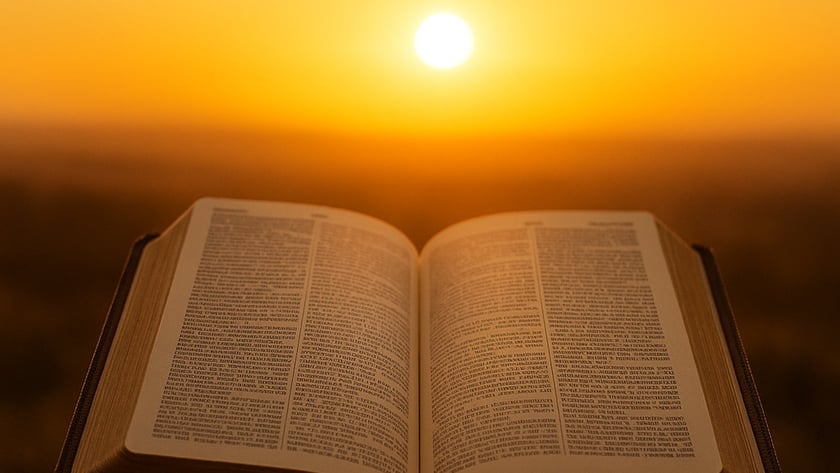Duas mãos seguram uma Bíblia aberta diante do sol se pondo no horizonte.