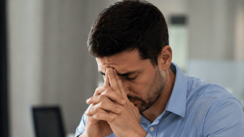 Homem de camisa azul apoia o rosto nas mãos diante de um notebook, em escritório.