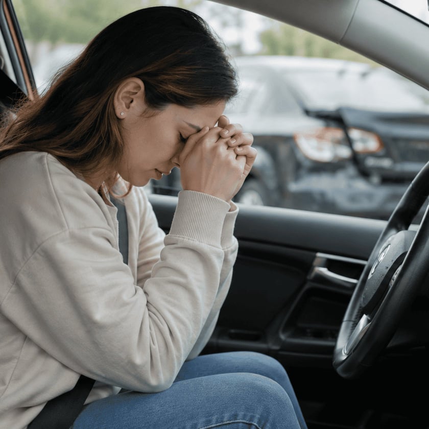Mulher sentada ao volante de um carro, com o rosto apoiado nas mãos unidas.