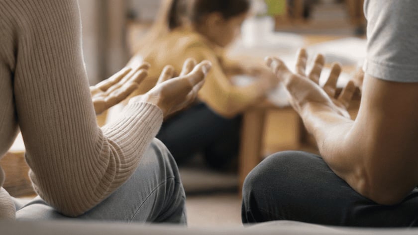 Duas pessoas sentadas num sofá com as palmas das mãos abertas; ao fundo, uma criança brinca.