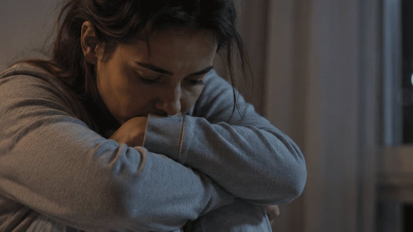 Mulher sentada na cama abraça os joelhos e apoia o rosto nos braços.