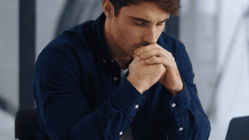 Homem de camisa azul com as mãos unidas junto ao rosto, diante de uma Bíblia aberta e um notebook.