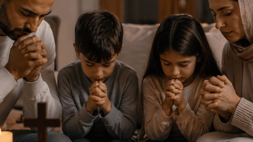 Homem, menino e menina rezam de mãos juntas diante de uma cruz, velas e Bíblia.
