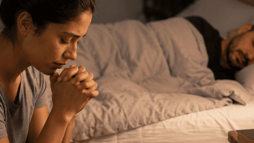 Homem dorme na cama enquanto uma pessoa, em primeiro plano, ora de mãos postas.