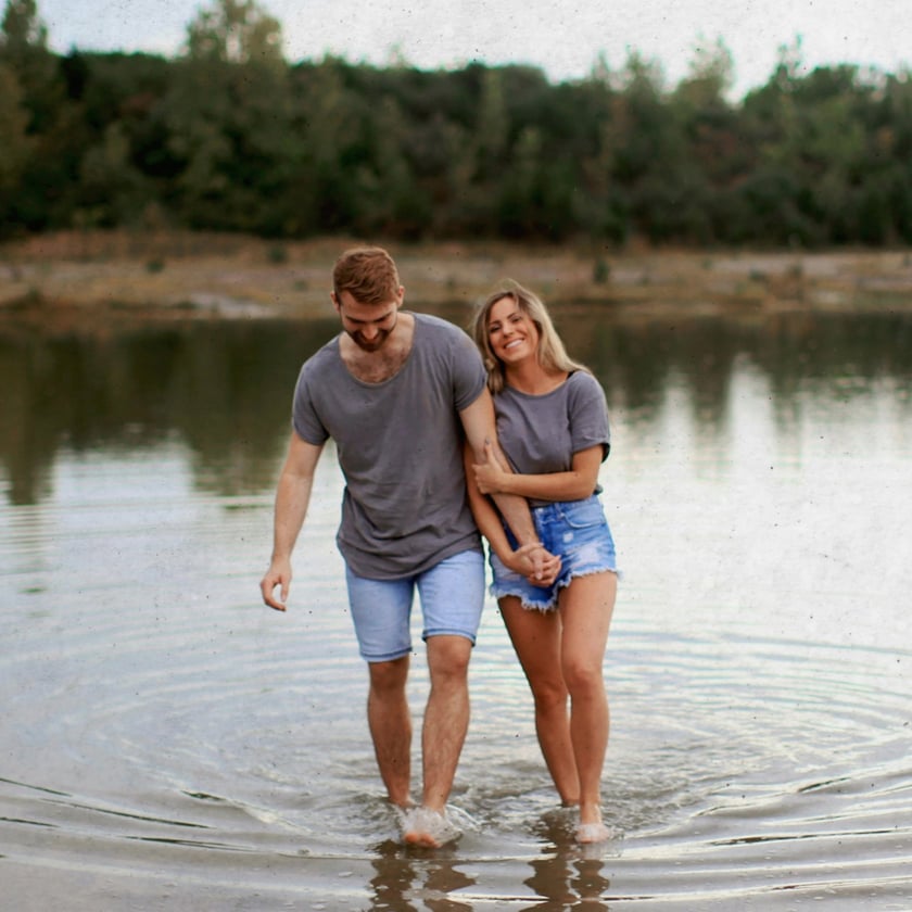 Casal de camisetas cinza e shorts jeans caminha de braços dados em um lago raso.