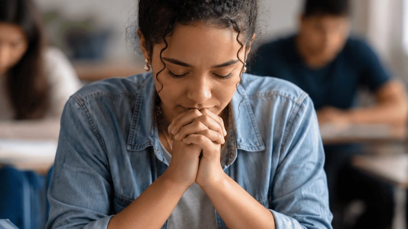 Jovem de cabelo cacheado e jaqueta jeans, de olhos fechados e mãos juntas, em sala de aula.