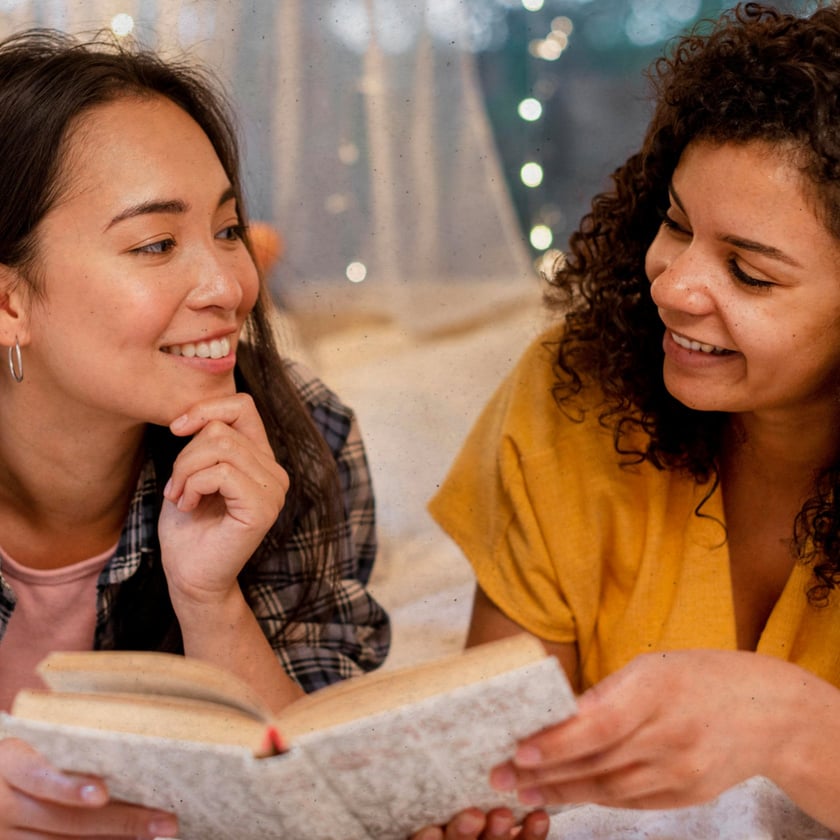 Duas mulheres sorridentes deitadas, olhando uma para a outra, com um livro aberto entre elas.