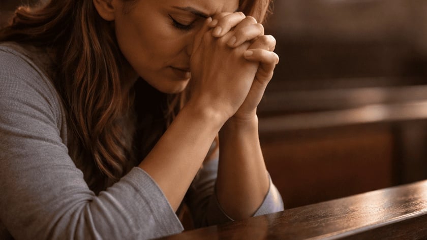 Mulher de cabelos castanhos com as mãos unidas junto à testa, orando em um banco de igreja.