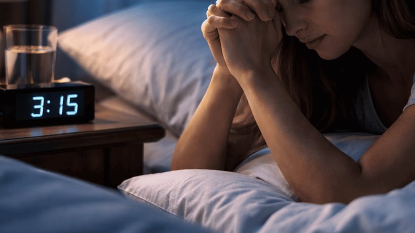 Mulher deitada na cama de mãos postas junto ao rosto, ao lado de um relógio marcando "3:15".