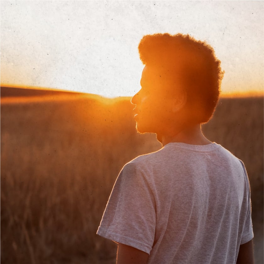 Jovem de camiseta clara, visto de costas e de perfil, olha um campo ao pôr do sol.